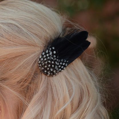 A close up of a blonde woman shown from the back wearing a black feather hair clip with white polka-dots, set against a blurred outdoor background.