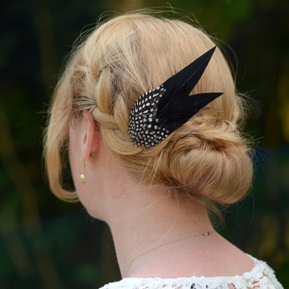 A blonde woman shown from the back wearing a black feather hair accessory with elegant sharp cut points and white polka dot spotted feathers in her low bun, set against a blurred dark green background.