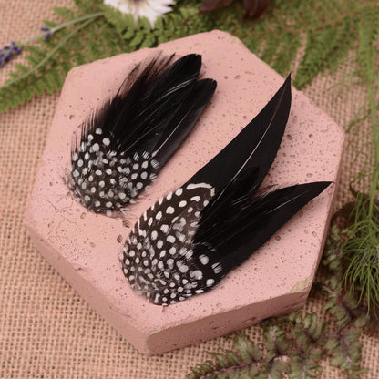 Two decorative black feather hair clips with white polka-dot spotted patterns, displayed on a pink hexagonal stone tile with burlap and green foliage in the background.