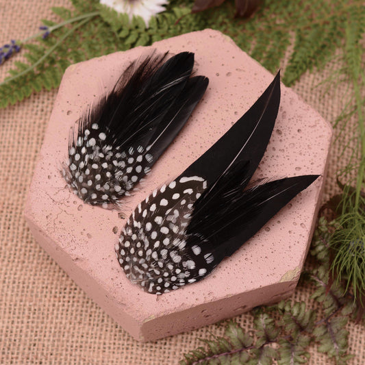 Two decorative black feather hair clips with white polka-dot spotted patterns, displayed on a pink hexagonal stone tile with burlap and green foliage in the background.