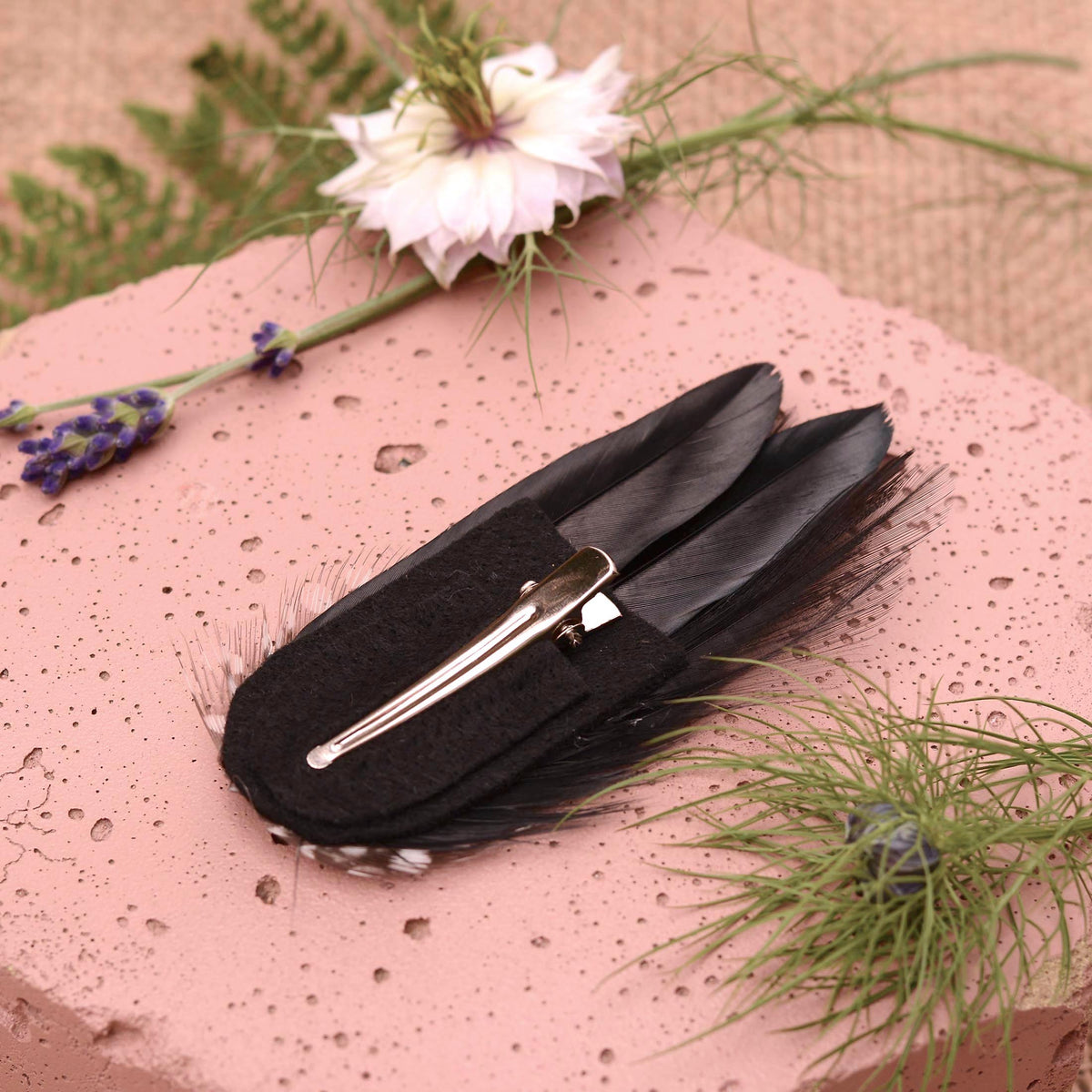 A black feather hair clip with a silver metal alligator-style clip and black felt backing , displayed on a textured pink stone with decorative flowers and greenery.