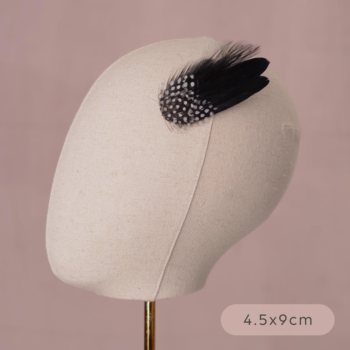 A black feather hair clip accessory with white spotted feathers, displayed on a beige fabric mannequin head against a dusty pink background, with a size label reading 4.5x9cm.