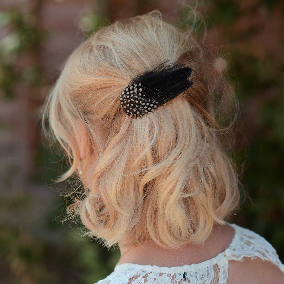 A blonde woman shown from the back wearing a black feather hair clip with white polka-dots, set against a blurred outdoor background.