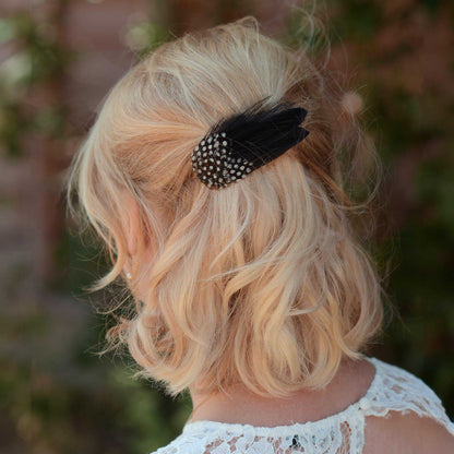 A blonde woman shown from the back wearing a black feather hair clip with white polka-dots, set against a blurred outdoor background.
