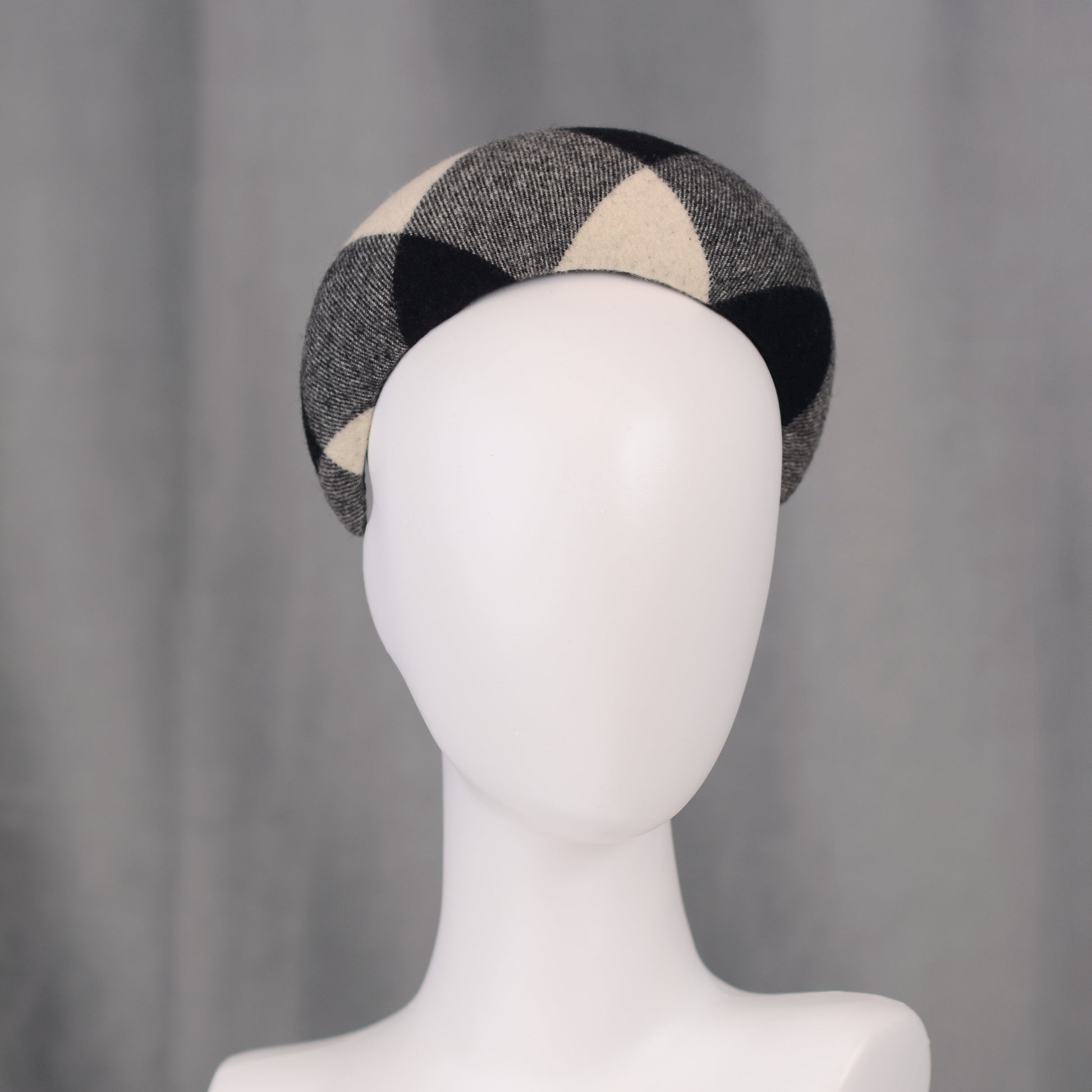 A black, white, and grey checked wool headband fascinator. The hat is displayed on a white mannequin head. The checked pattern is made up of large squares.