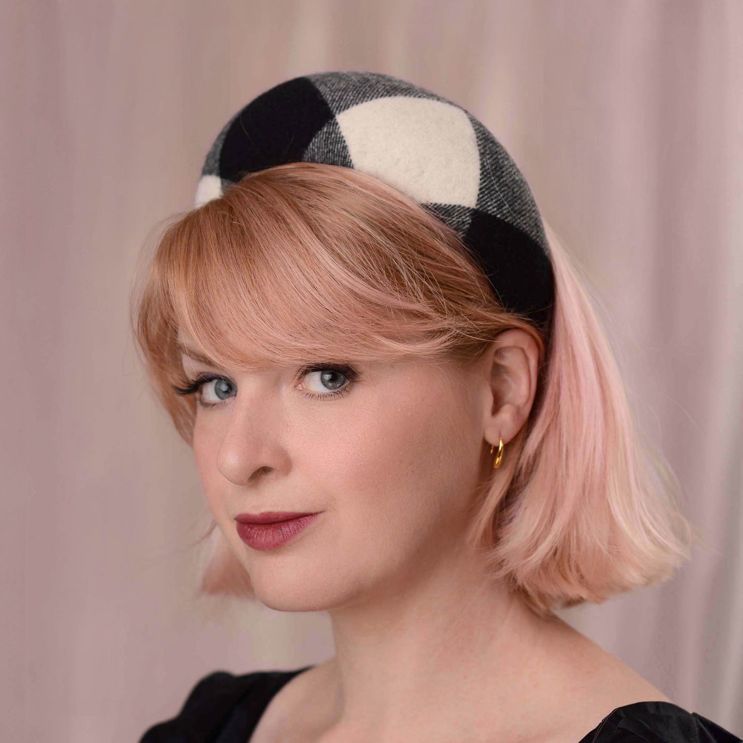 A woman with short pink hair wearing a black and ivory checkered wool headband, photographed against a soft pink curtain background.