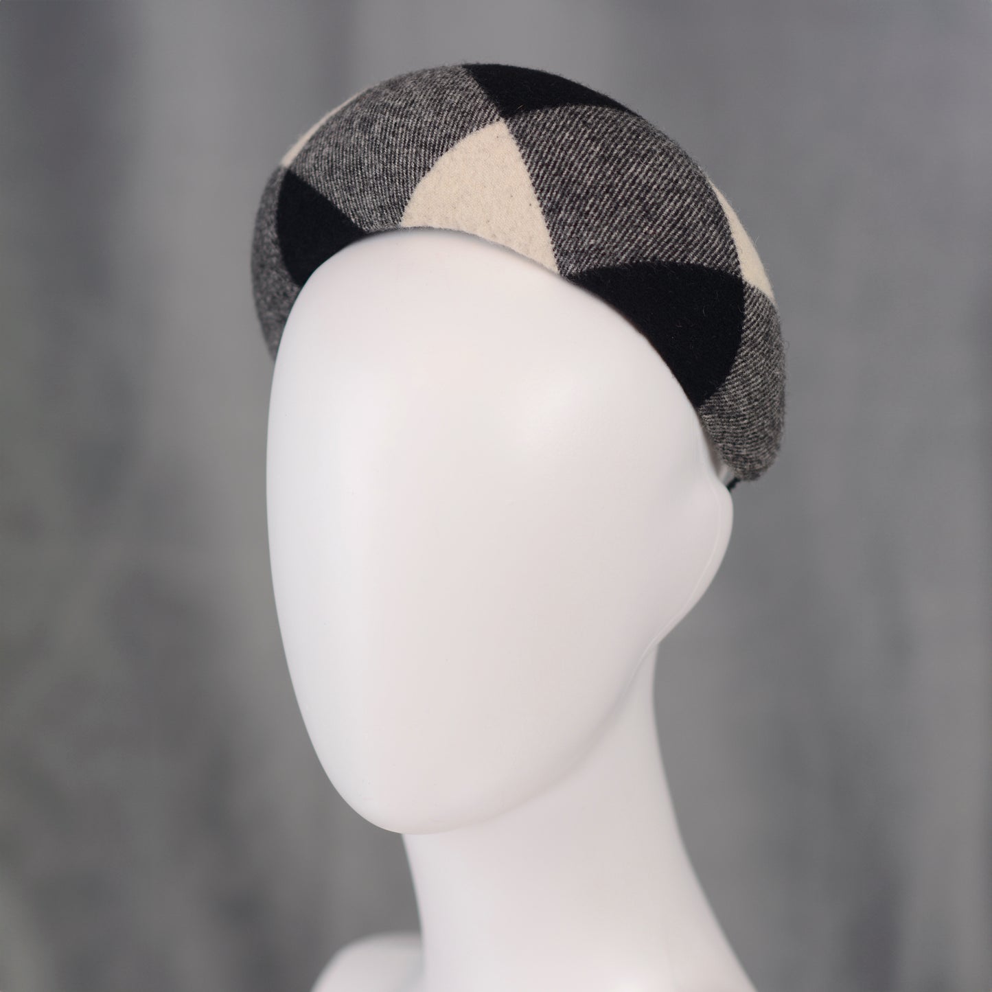A black, white, and grey checked wool headband fascinator. The hat is displayed on a white mannequin head. The checked pattern is made up of large squares.