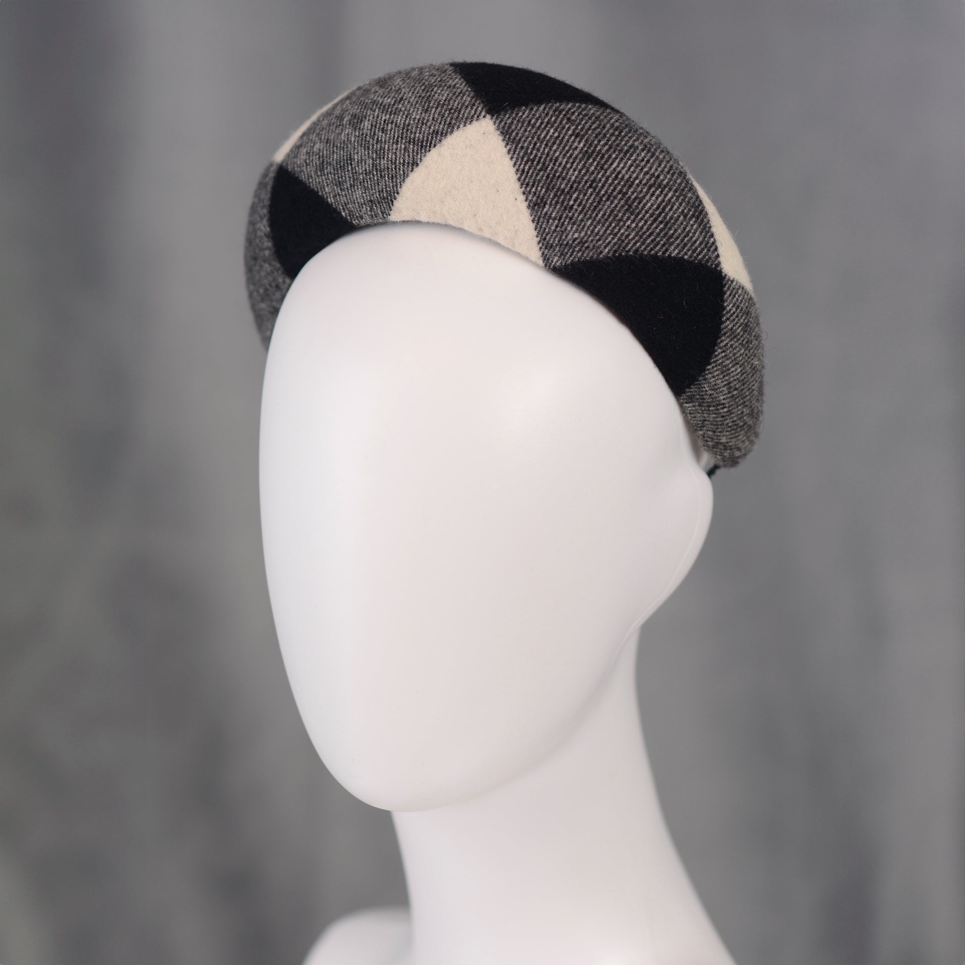A black, white, and grey checked wool headband fascinator. The hat is displayed on a white mannequin head. The checked pattern is made up of large squares.