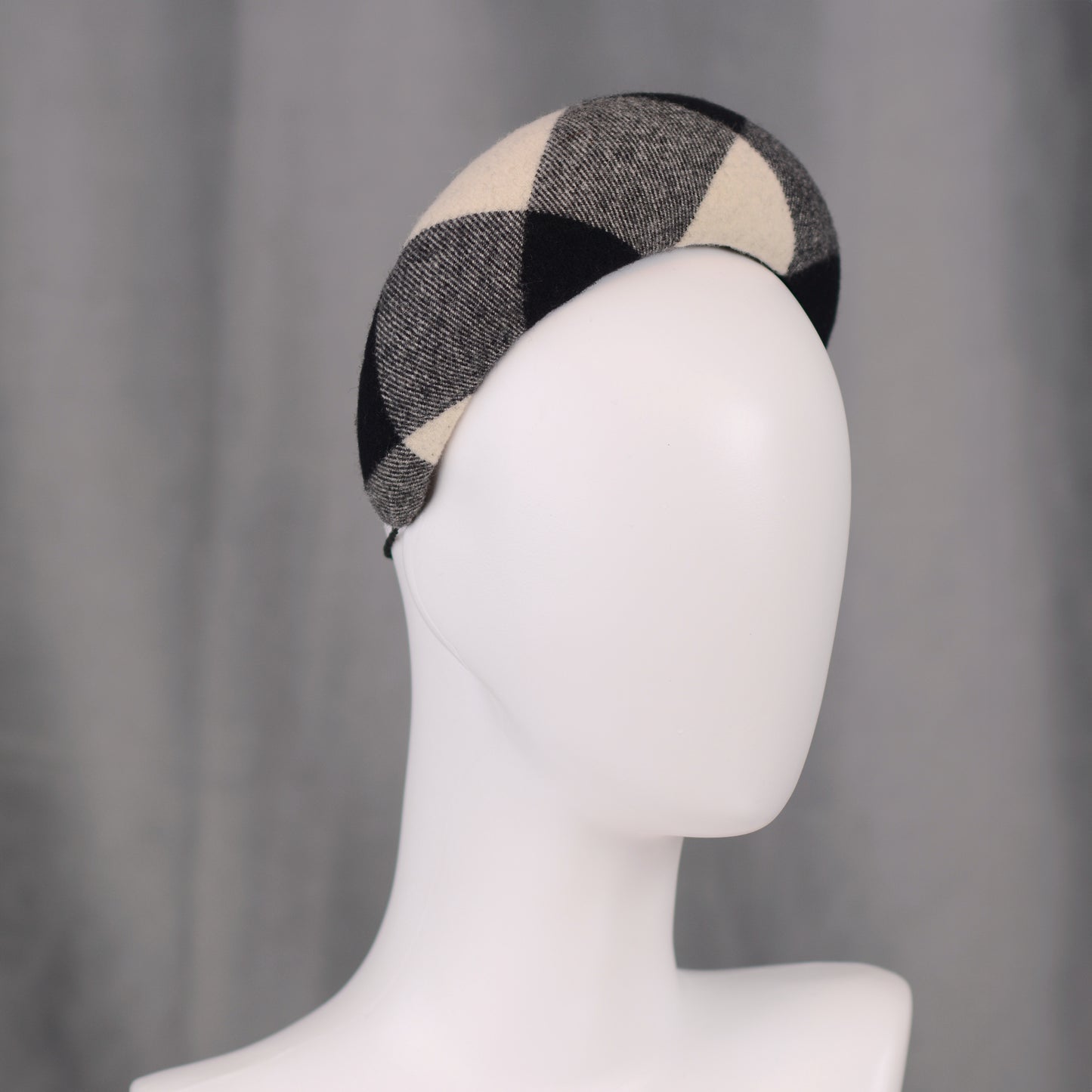 A black, white, and grey checked wool headband fascinator. The hat is displayed on a white mannequin head. The checked pattern is made up of large squares.