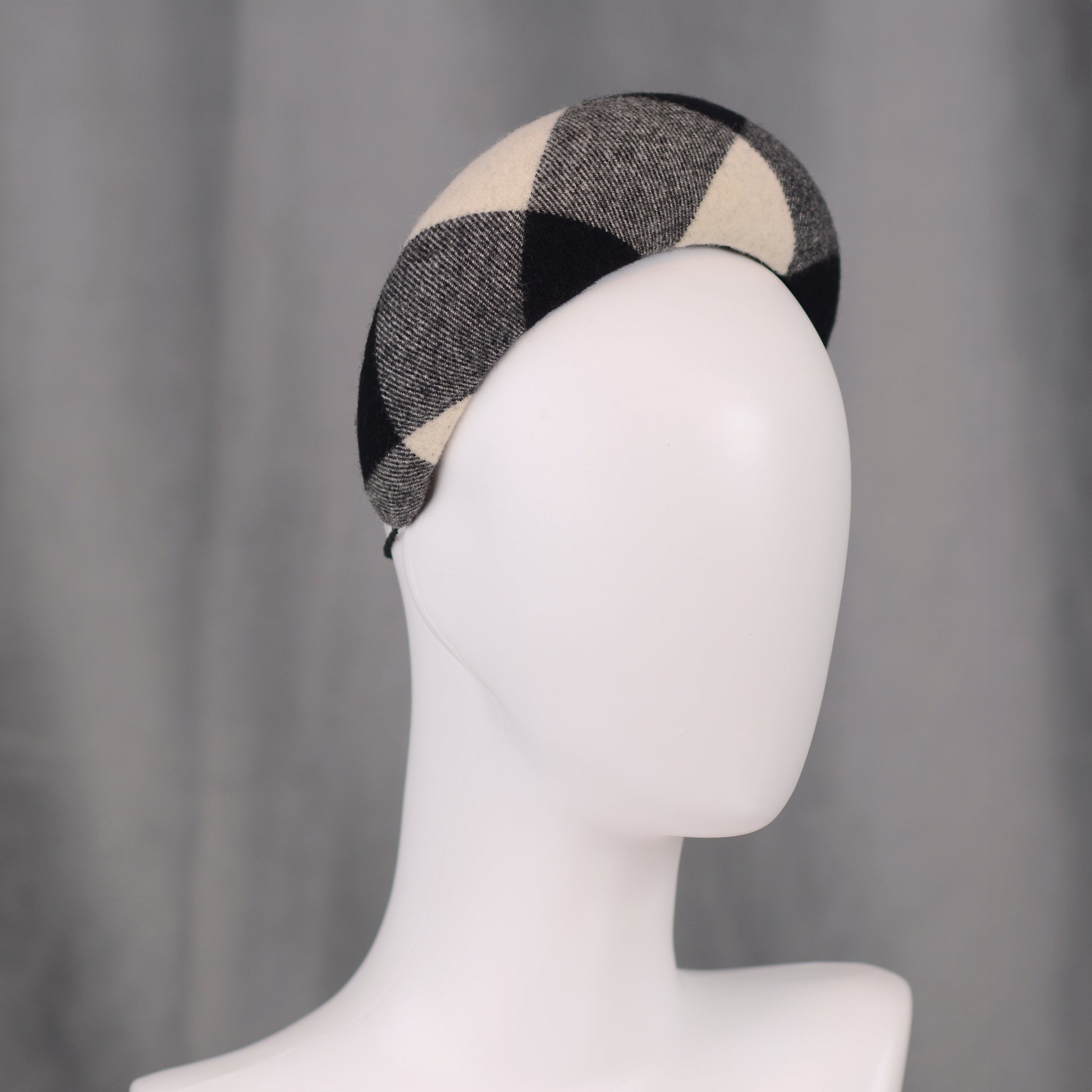 A black, white, and grey checked wool headband fascinator. The hat is displayed on a white mannequin head. The checked pattern is made up of large squares.