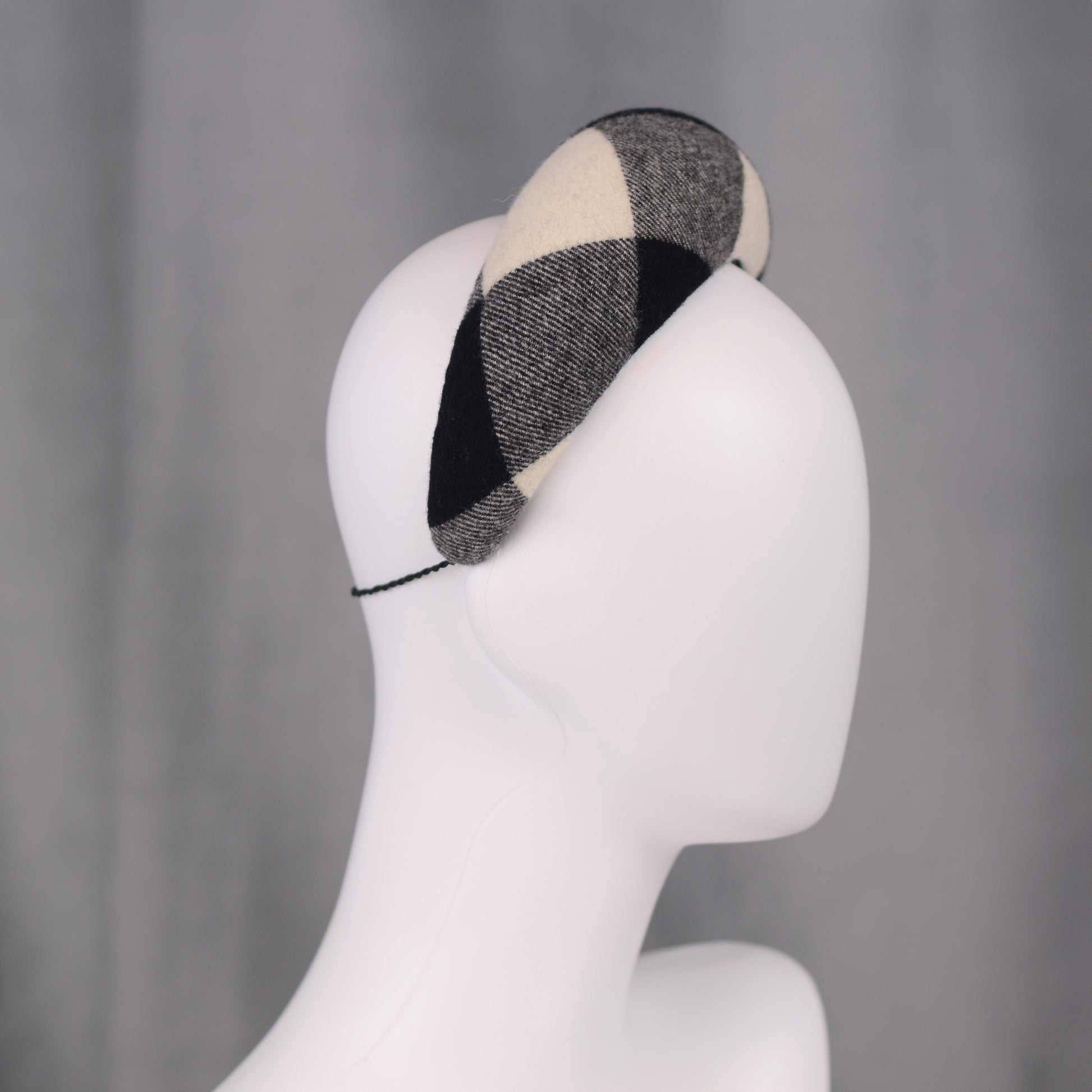 A black, white, and grey checked wool headband fascinator. The hat is displayed on a white mannequin head. The checked pattern is made up of large squares.