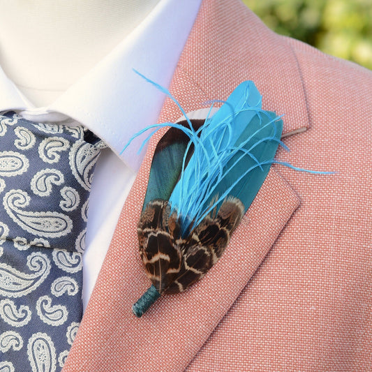 A close-up of a men's formal wear ensemble featuring a salmon pink textured suit jacket, a white dress shirt, a blue and white paisley patterned tie, and a vibrant turquoise blue feather boutonniere pin on the lapel.