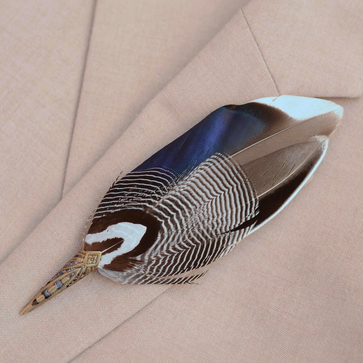 A decorative duck feather brooch pin with grey  white, and iridescent blue colouring, attached to a antique brass toned metal cap, displayed on a beige textured fabric background.