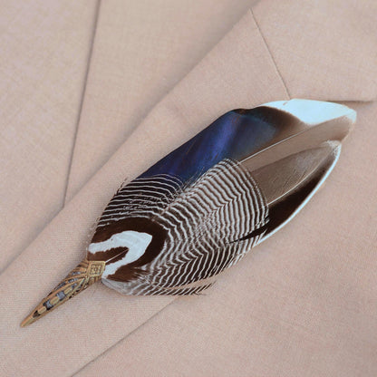 A decorative duck feather brooch pin with grey  white, and iridescent blue colouring, attached to a antique brass toned metal cap, displayed on a beige textured fabric background.