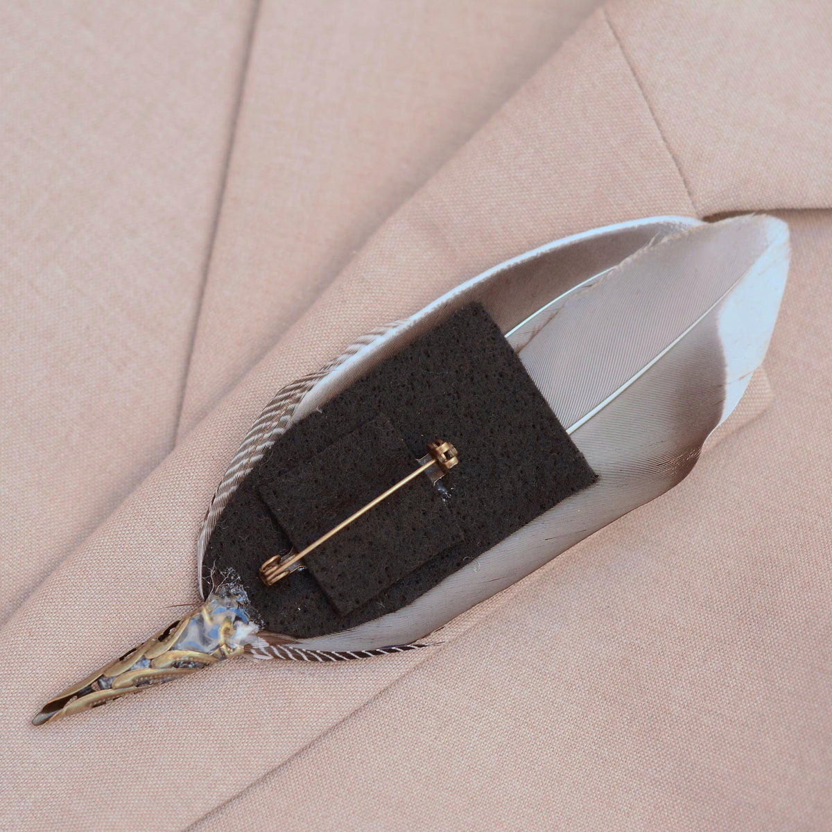 A decorative bird feather pin with a antique brass-toned metal cap and brooch pin on a felt base and resting on a light beige textured fabric background.