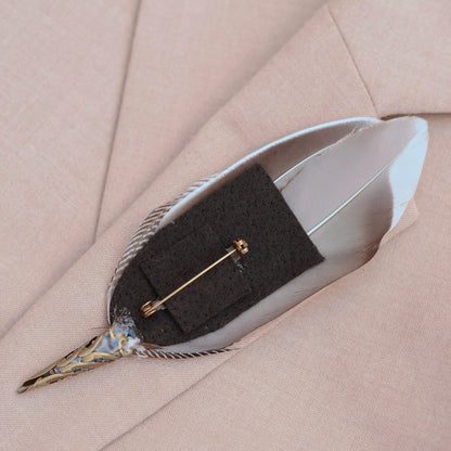 A decorative bird feather pin with a antique brass-toned metal cap and brooch pin on a felt base and resting on a light beige textured fabric background.