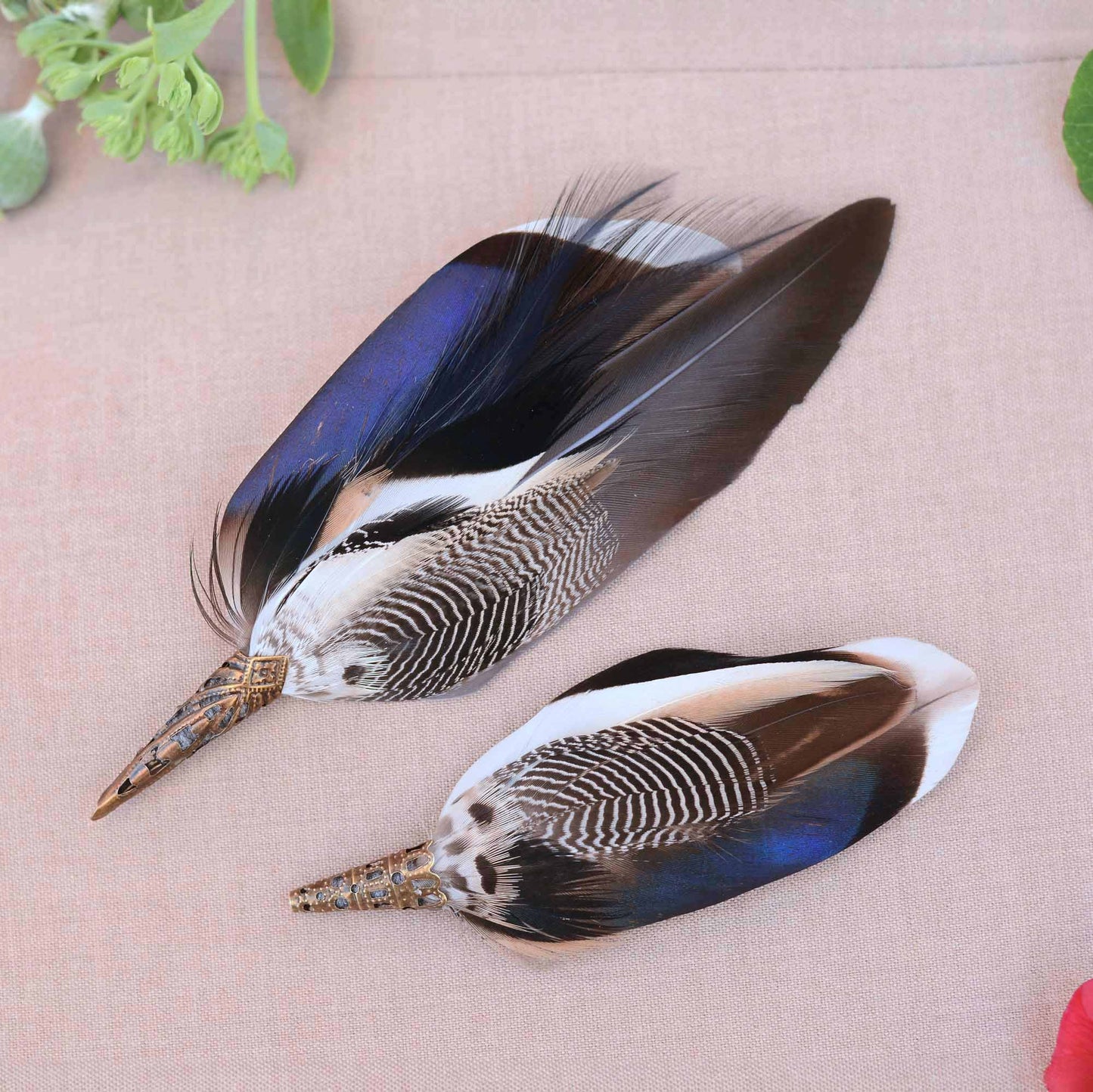 Two decorative duck feather broches with iridescent blue, black, white, and brown tones, laid on a light beige textured surface with small green leaves at the corners.