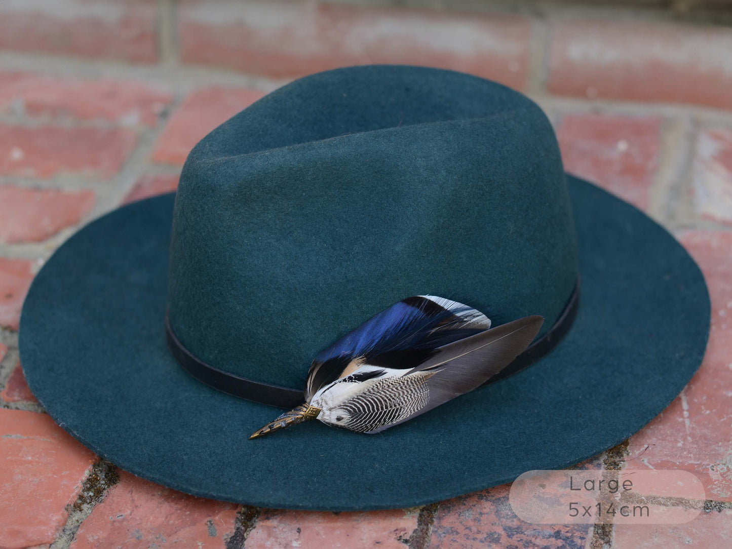 A dark teal felt fedora hat with a black band and a decorative duck feather hat pin in iridescent blue, grey and black, resting on a red brick surface.