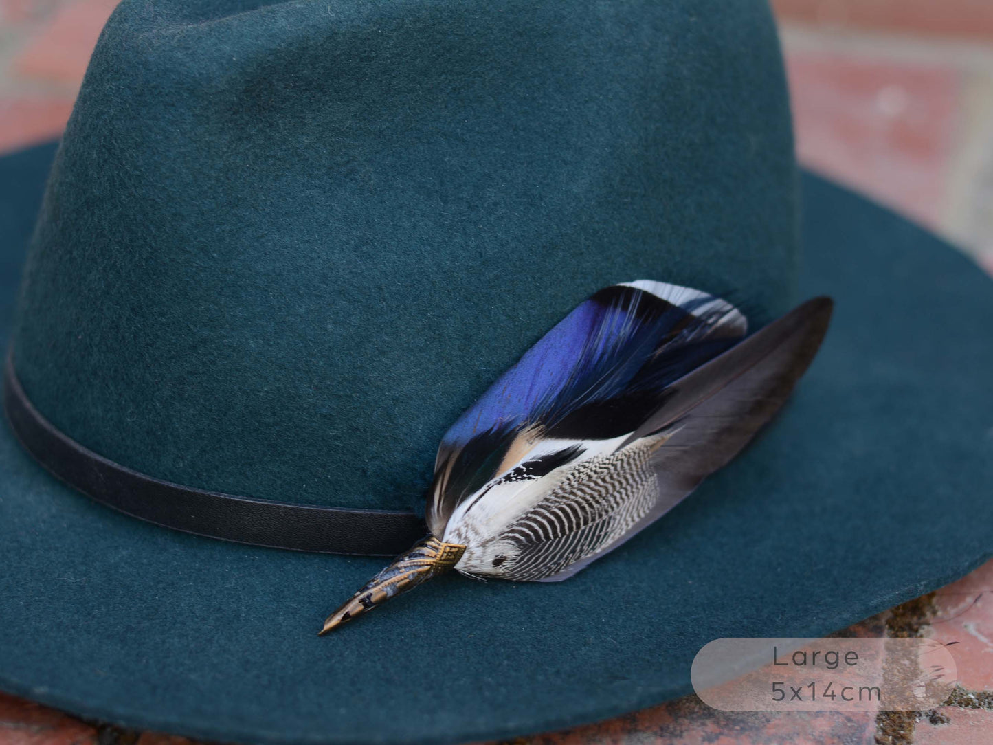 Close up of a dark teal felt fedora hat with a black band and a large decorative duck feather hat pin in iridescent blue, grey and black, resting on a red brick surface.