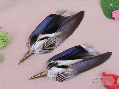 Two decorative duck feathers brooches with patterned brown, black, white, and iridescent blue tops, attached to metal pins with white backing paper, displayed on a beige fabric background with small floral props.