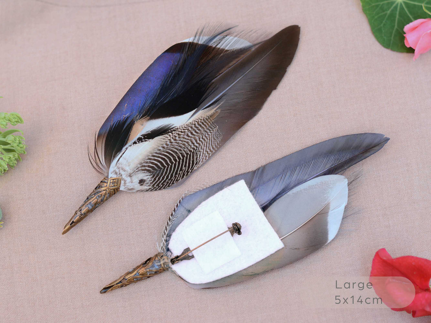 Two decorative duck feathers brooches with patterned brown, black, white, and iridescent blue tops, attached to metal pins with white backing paper, displayed on a beige fabric background with small floral props.
