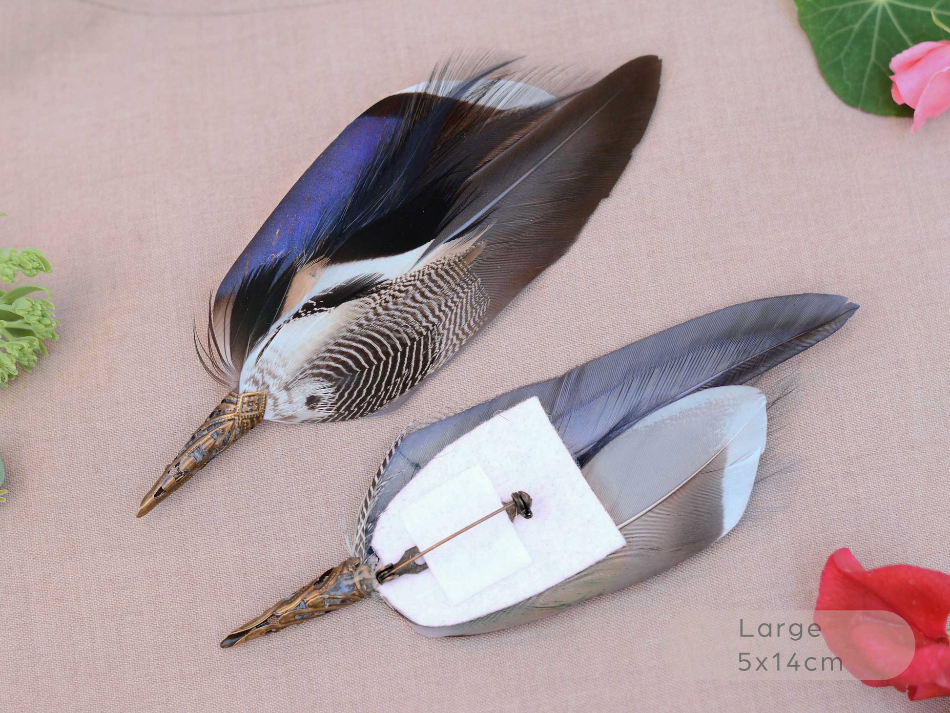 Two decorative duck feathers brooches with patterned brown, black, white, and iridescent blue tops, attached to metal pins with white backing paper, displayed on a beige fabric background with small floral props.