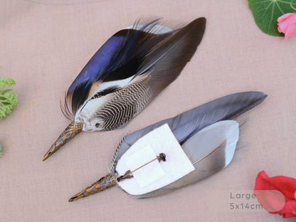 Two decorative duck feathers brooches with patterned brown, black, white, and iridescent blue tops, attached to metal pins with white backing paper, displayed on a beige fabric background with small floral props.