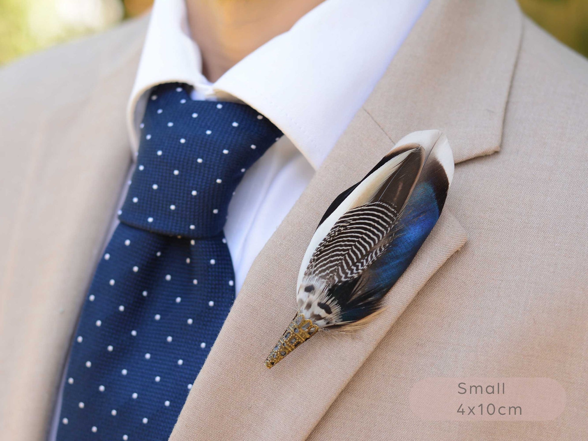 A close-up of a beige suit jacket worn with a white dress shirt, navy polka-dot tie, and a decorative duck feather lapel pin. The pin has black-and-white striped detailing with blue, black, and white feathers.