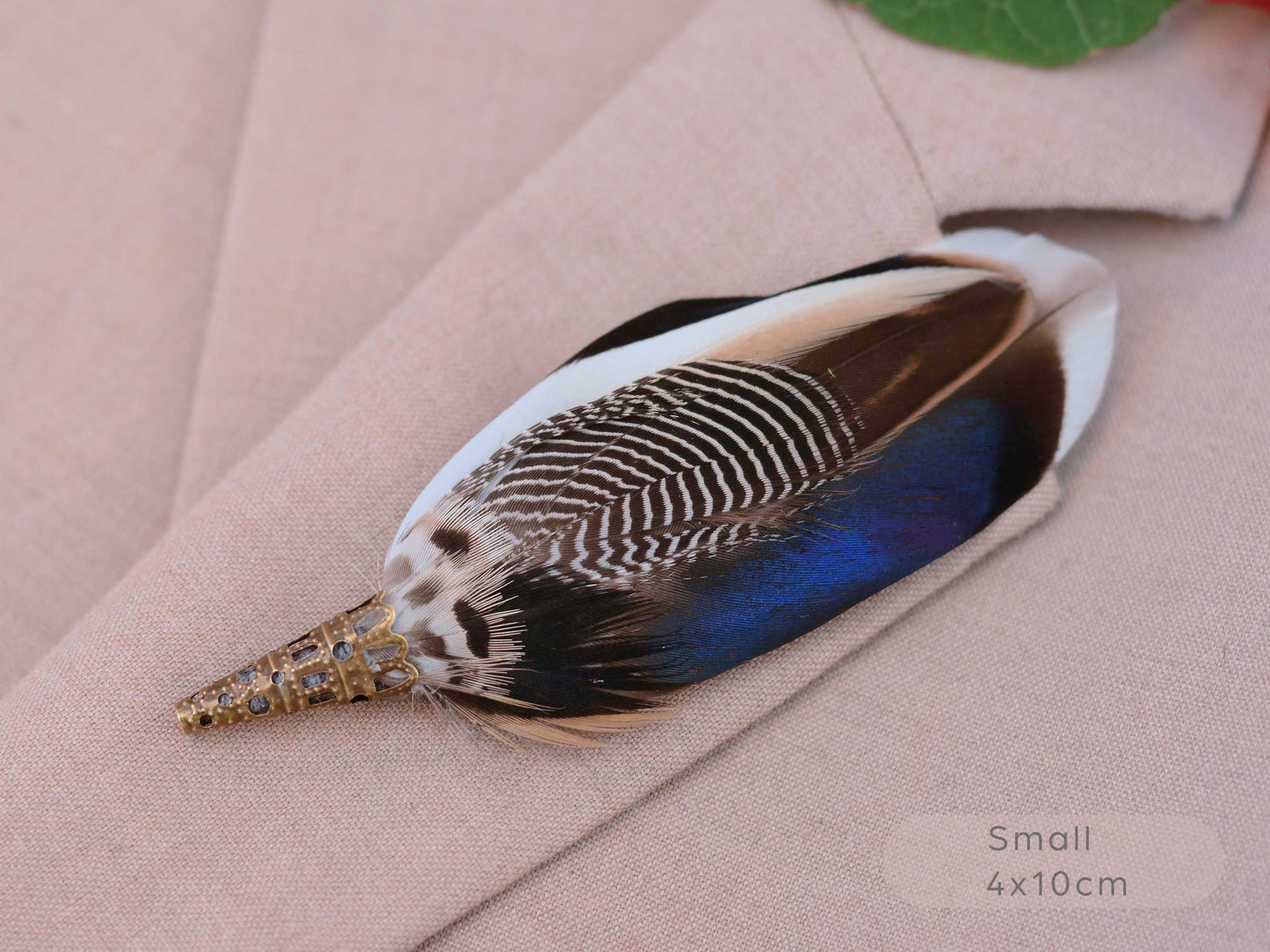 A decorative feather pin with a brass-toned conical base and a large patterned feather in white, brown, black, and vivid blue, displayed on a beige fabric surface. Text in the corner reads 'Small 4x10cm'.