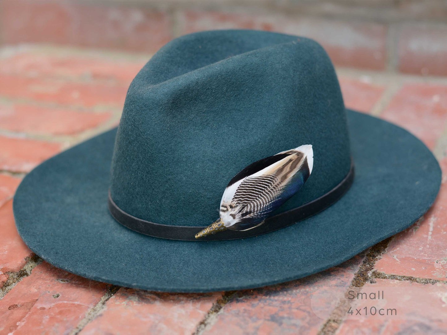 A dark teal felt fedora hat with a black band and a decorative duck feather hat pin, resting on a red brick surface.