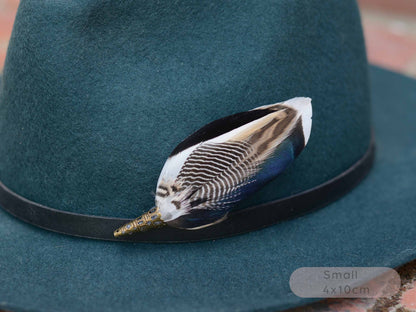 A close-up of a dark teal felt hat with a black band, decorated with a small bird feather pin featuring black, white, blue, and brown patterned plumage with a gold-toned beak. A small 'Small 4x10cm' label is visible in the bottom right corner.
