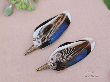 Two decorative bird feather buttonholes with iridescent blue, brown, black, and white patterns, displayed on a textured beige fabric background with a small green leaf in the corner and 'Small 4x10cm' text in the bottom right.