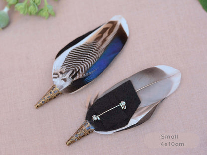 Two decorative bird feather buttonholes with iridescent blue, brown, black, and white patterns, displayed on a textured beige fabric background with a small green leaf in the corner and 'Small 4x10cm' text in the bottom right.