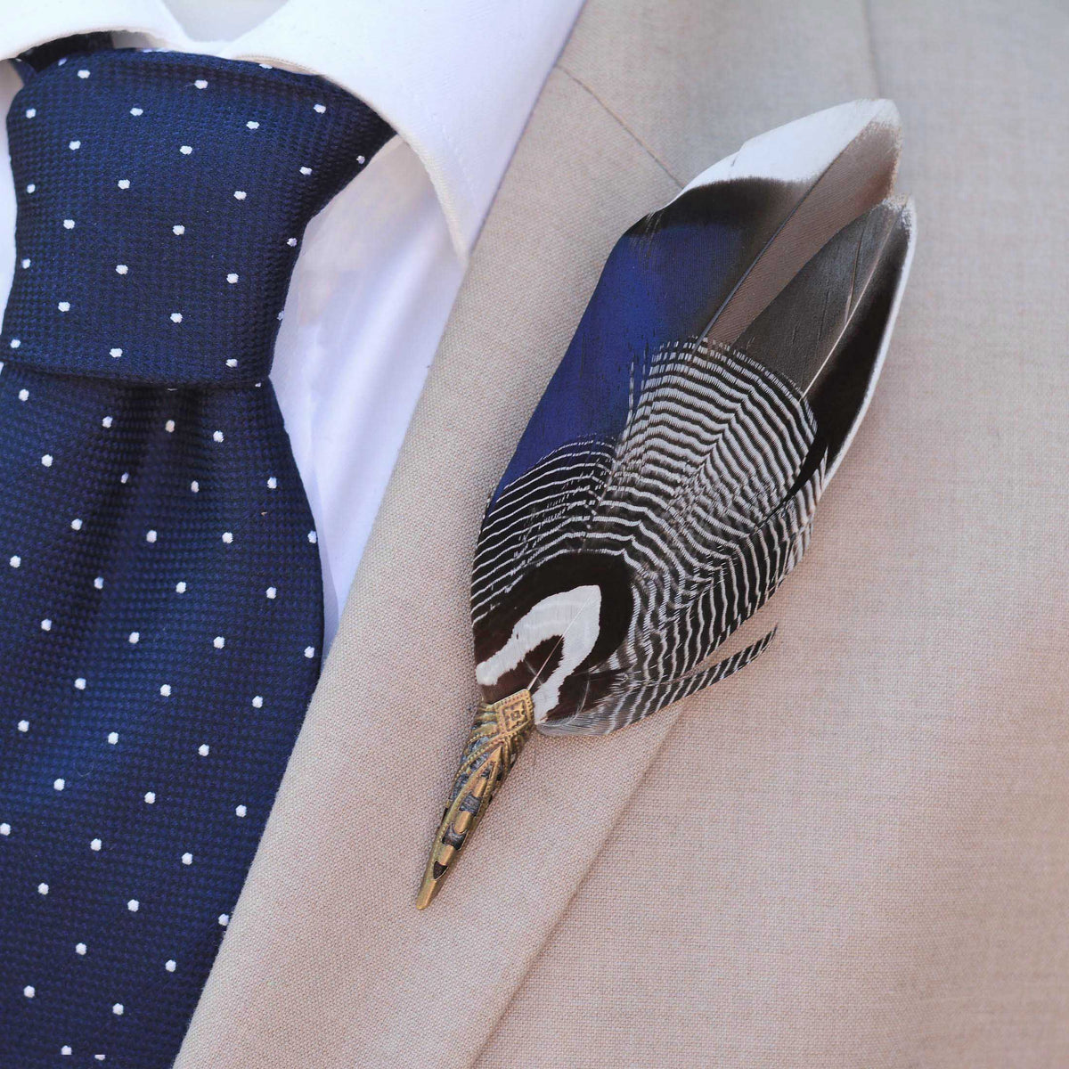 A decorative bird feather boutonniere pin with black, white, and blue patterned feathers and a brass toned metal cap, pinned to a beige suit jacket worn with a navy polka dot tie and white dress shirt.