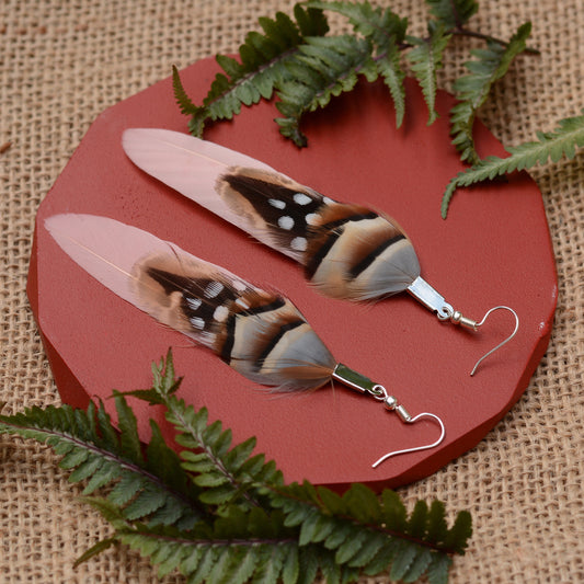 A pair of dangling earrings made from long feathers with brown, black, white, and pale pink tones, attached to silver fishhook wires, displayed on a round reddish platform with burlap and green ferns around them.