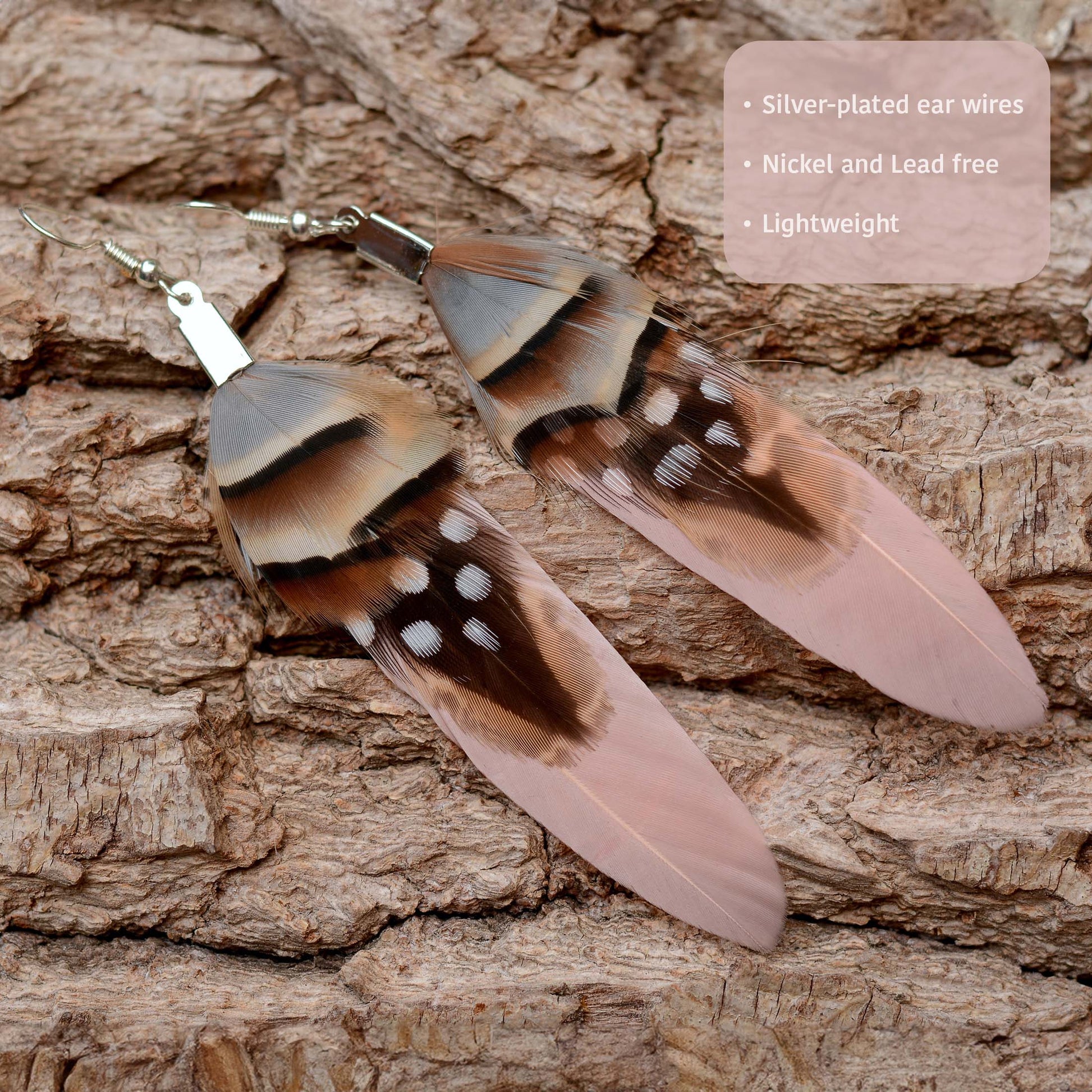 A pair of long feather statement earrings with brown and black bands, white spots, and pale pink tips, displayed on a textured brown bark background.