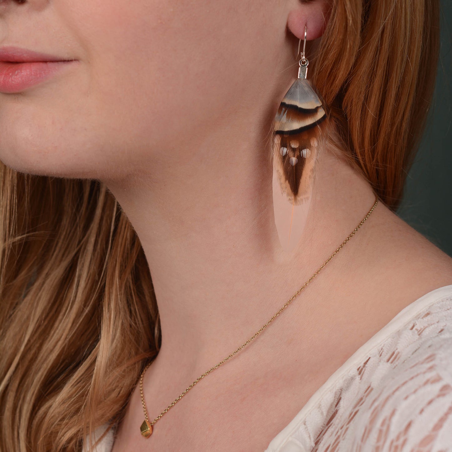 A close up of a woman with long blonde hair wearing a long feather drop earring of layered real feathers with soft brown and blue tones and strong markings on a soft pink duck wing feather and silver plated fish hook ear wires.