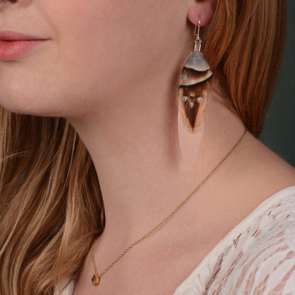 A close up of a woman with long blonde hair wearing a long feather drop earring of layered real feathers with soft brown and blue tones and strong markings on a soft pink duck wing feather and silver plated fish hook ear wires.