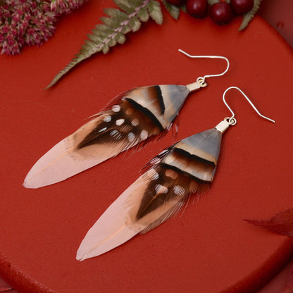 A pair of soft pink  feather drop earrings with pheasant, guinea fowl and partridge feathers and silver plated fishhook wires, displayed on a textured red surface with decorative dried flowers and green leaves in the background.