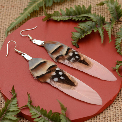 A pair of long feather drop festival earrings with silver fishhook wires, displayed on a round red plate with green fern leaves on a burlap background.