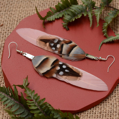 A pair of long  real feather earrings made from patterend natural feathers with silver fishhook wires, displayed on a red circular stand with burlap and fern leaves.