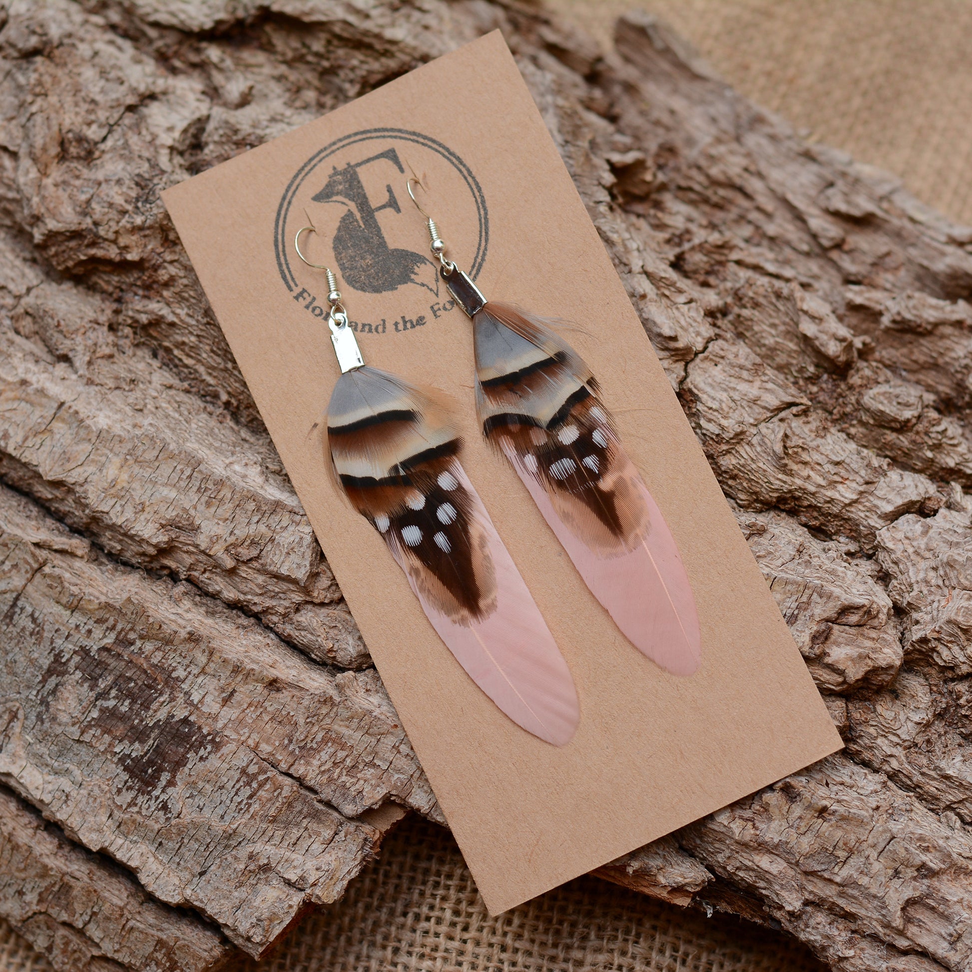 A pair of dangling  real feather earrings displayed on a kraft card, placed on textured tree bark. The earrings have light pink feathers with dark brown and black bands, white spots, and silver-plated hooks.