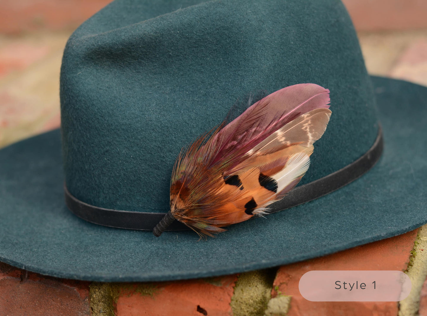 Green hat with a large burgundy and pheasant feather hat pin on a textured brick surface