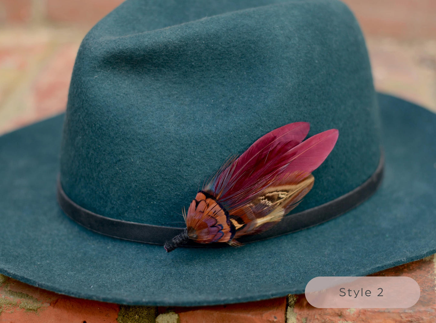 Green hat with a small burgundy and pheasant feather hat pin on a textured brick surface