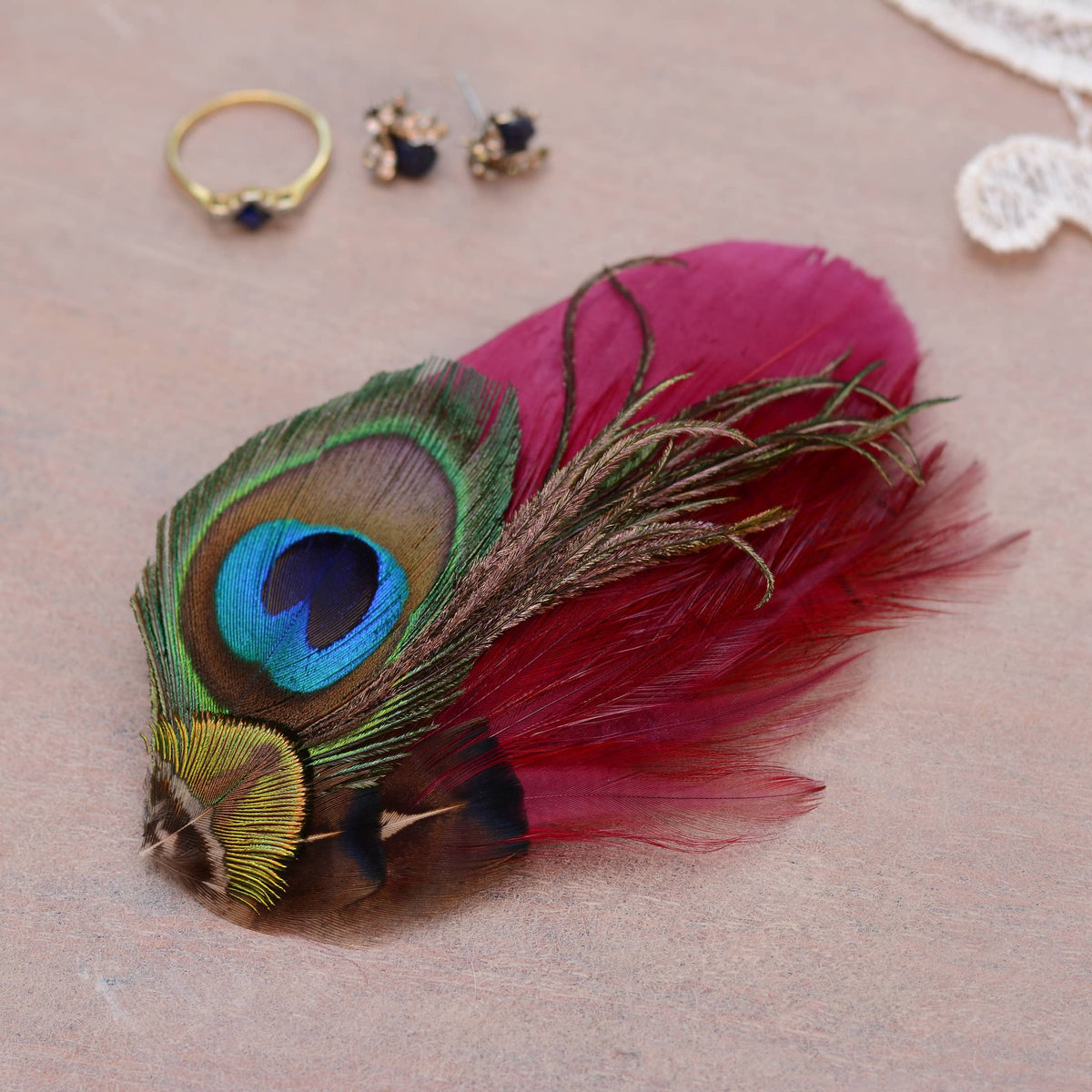 A decorative feather hair accessory featuring a peacock feather with a vivid blue and green eye pattern layered on top of burgundy feathers displayed on a neutral beige surface with small jewellery props nearby.