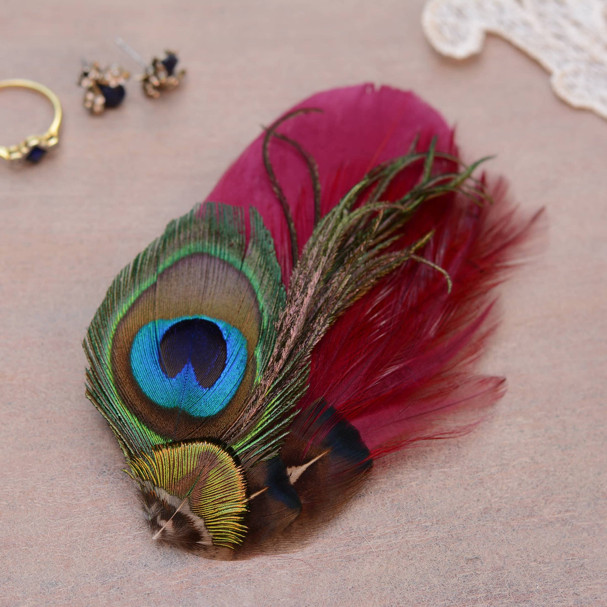 A decorative feather accessory featuring a peacock feather with a vivid blue, green, and brown eye pattern, paired with a lred burgundy feathers, displayed on a neutral beige surface with small jewelry props in the background.