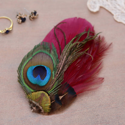 A decorative feather accessory featuring a peacock feather with a vivid blue, green, and brown eye pattern, paired with a lred burgundy feathers, displayed on a neutral beige surface with small jewelry props in the background.