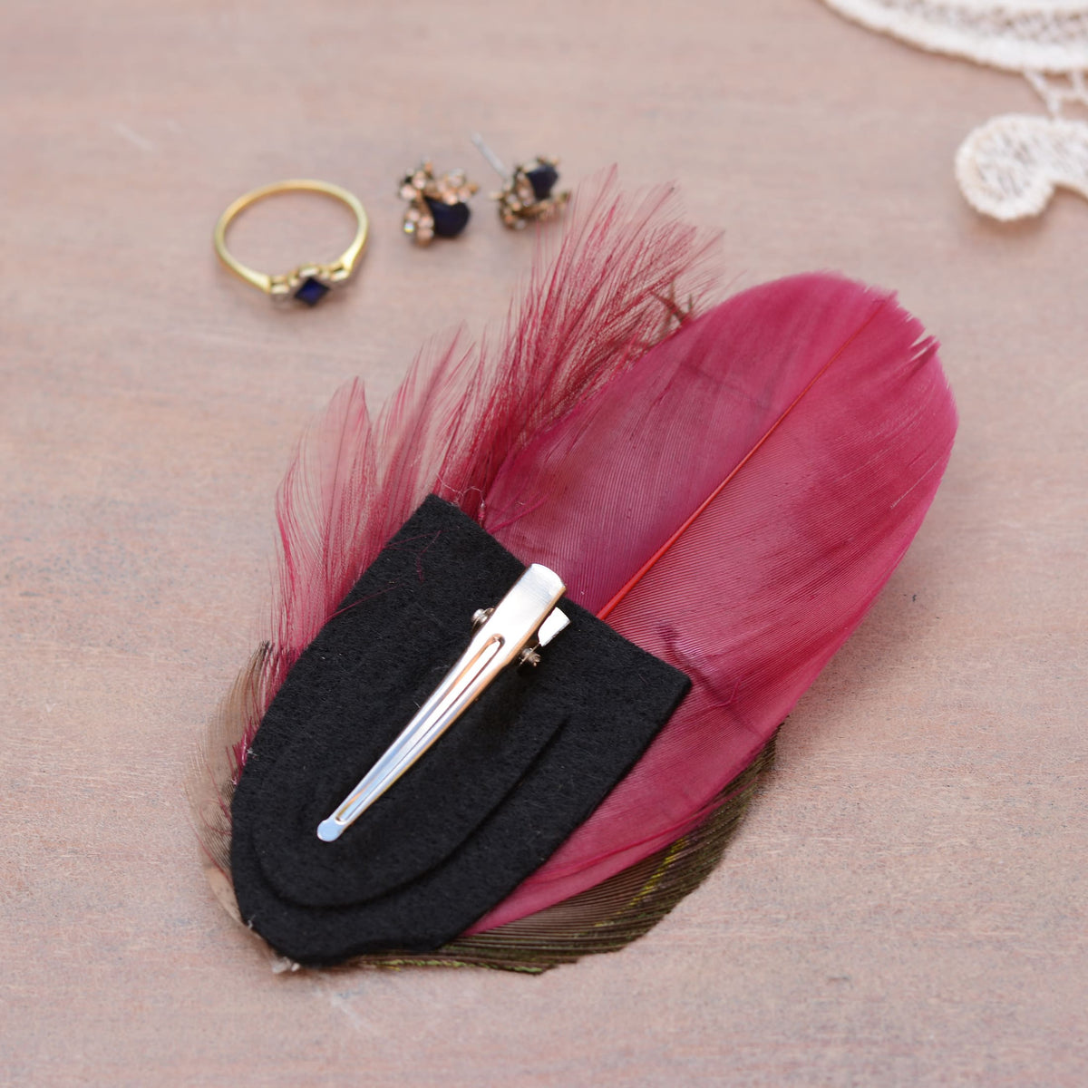 A decorative hair clip featuring peacock and burgundy feathers on a black felt backing with a silver metal crocodile clip, styled on a neutral textured surface with small jewellery props