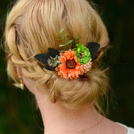 Blonde hair styled in an updo with decorative flower hair clip in burnt orange and green against a blurred natural background 