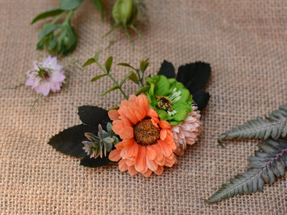 An artificial floral hair accessory featuring a prominent orange daisy with a brown center, a light pink flower, a green flower, and dark green leaves, styled on a textured beige burlap background.